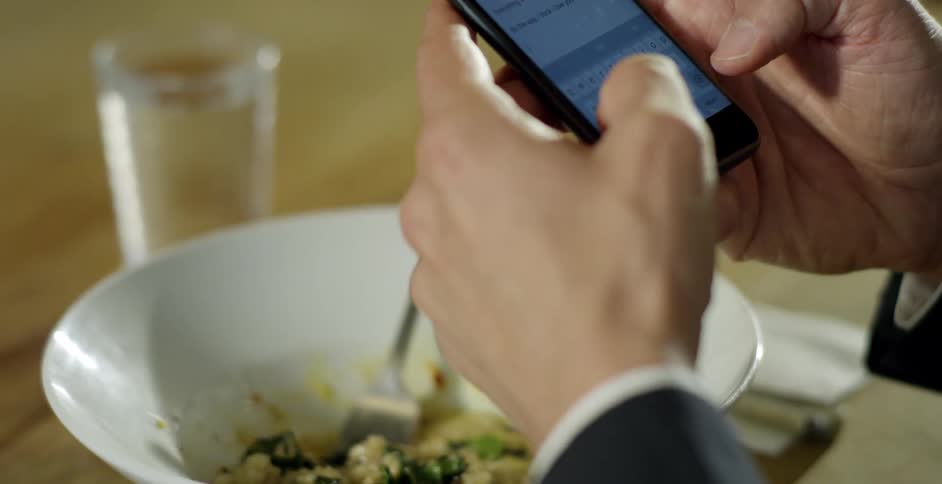 A man types on a smartphone while sitting in front of his food at a dining table