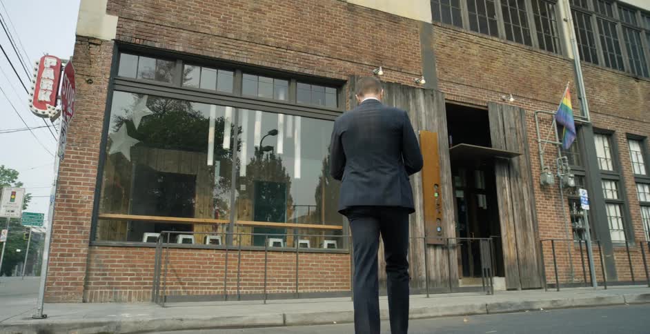 A man in a suit and sunglasses crosses a city street as he walks towards the entrance of a building