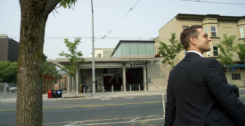 A man smiles as he walks down a city street in a black suit and sunglasses