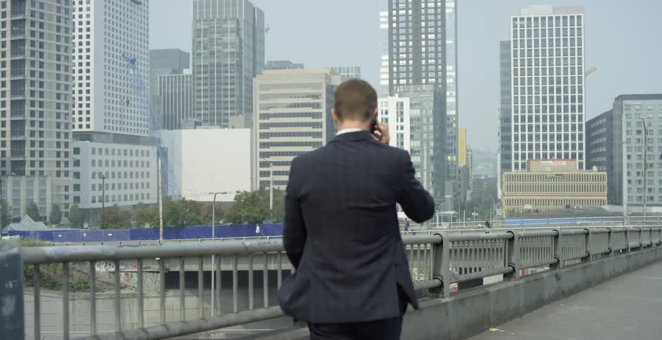A man in suit walks across bridge in a metropolis while holding a smartphone to his ear