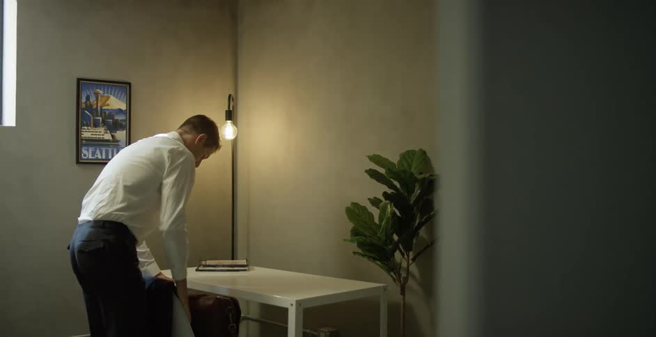 A man gathers his laptop from his desk and places it in his satchel before leaving his office