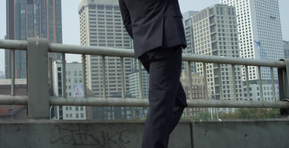 A man in a suit and sunglasses walks across a bridge in metropolis with his smartphone held to his ear