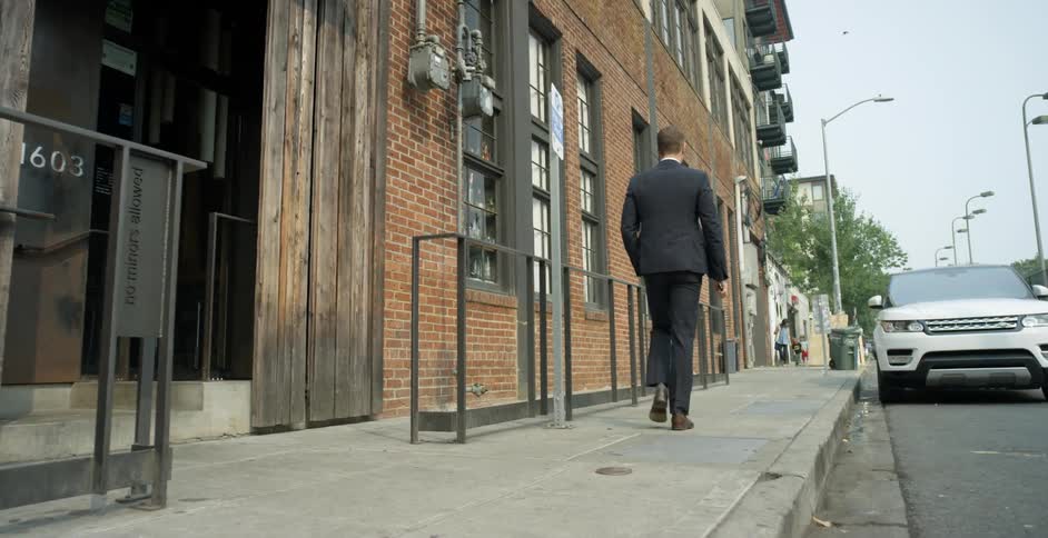 Man with a black suit and a pair of formal brown shoes holds a smartphone and walks down a street