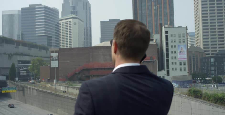 Man in a suit and sunglasses talks on the phone in front of the citys downtown skyscrapers