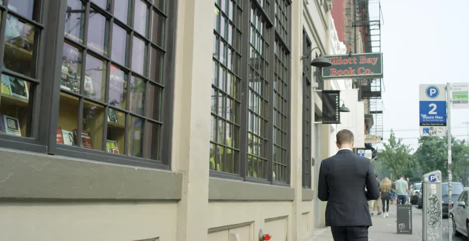 Man in a suit walks on the sidewalk of a retail section of the city as cars are parked on the street