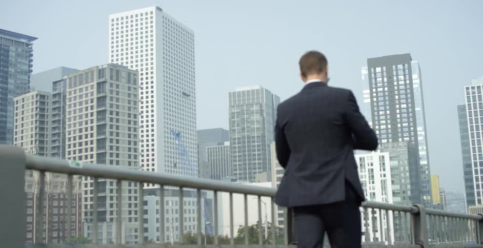 A man with a cell phone and a suit walking next to handrailing with buildings and the sky in the background
