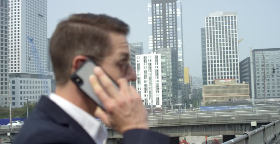 A man stands on a sidewalk in a large city talking on a cell phone