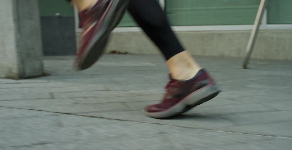 A persons feet with sneakers running on the pavement with buildings and windows in the background