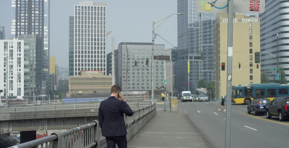A man in a suit and sunglasses walks down a sidewalk next to a busy road while holding his smartphone to his ear