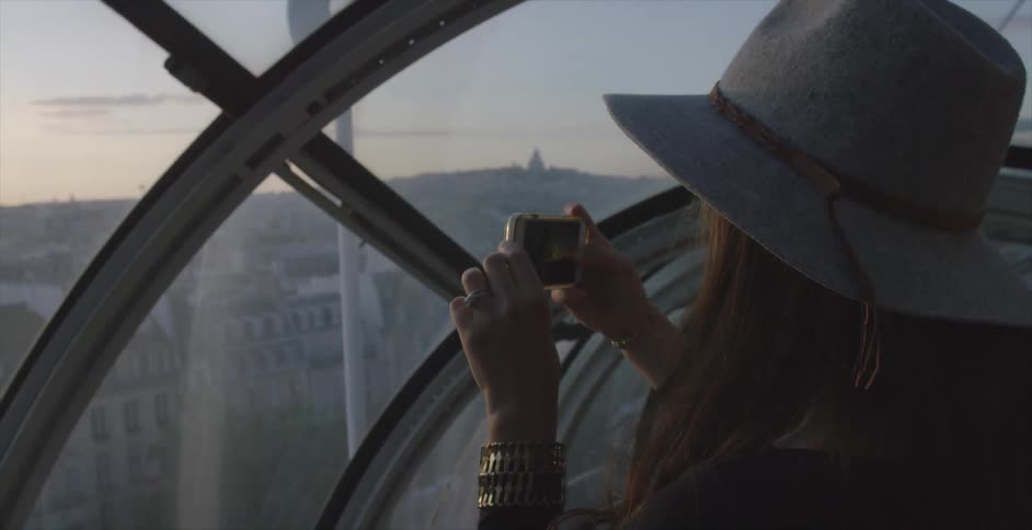 A woman is using her phone to photograph a city from a window at sunset