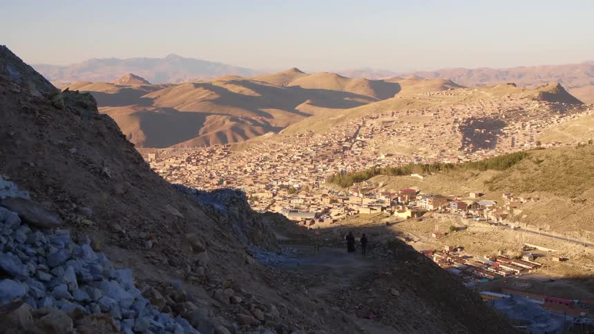 People and dog walk along slope of mountain with town occupied valley in background