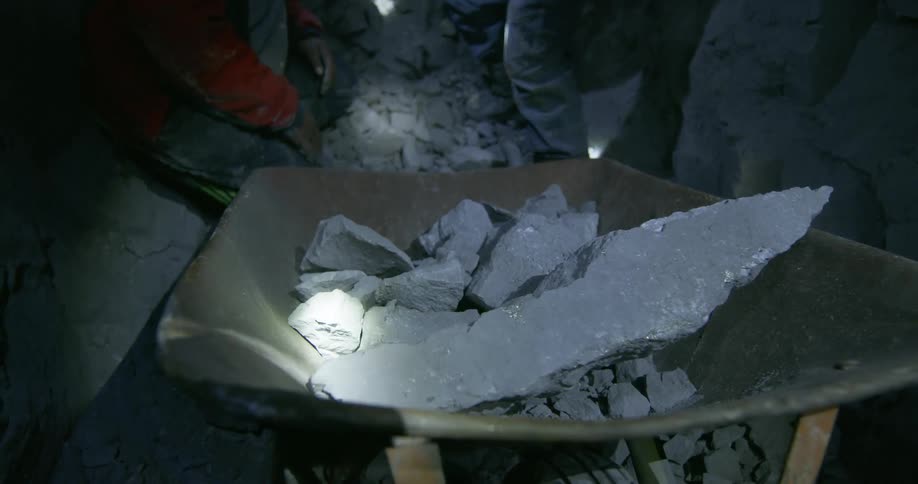 A miner tosses a rock into a wheelbarrow in a dark mine