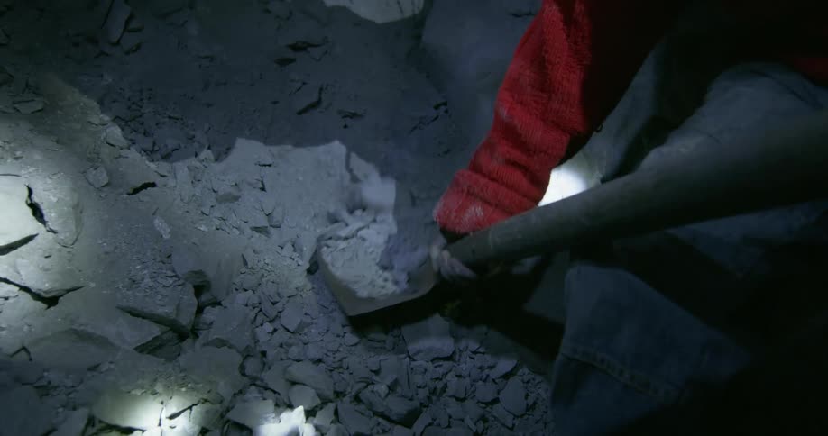 A miner shovels dirt and rocks into a wheelbarrow in a dark mine