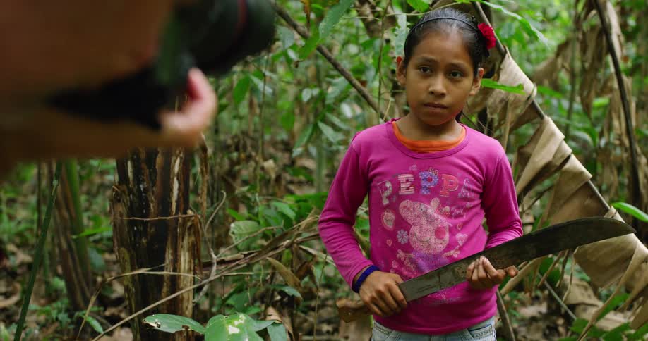A young girl stands in the jungle holding a machete with an adult taking photos