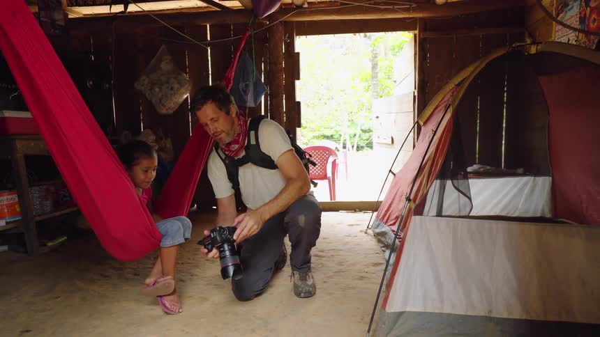 Photographer shows picture on his camera to a girl sitting in a hammock inside a wooden shack