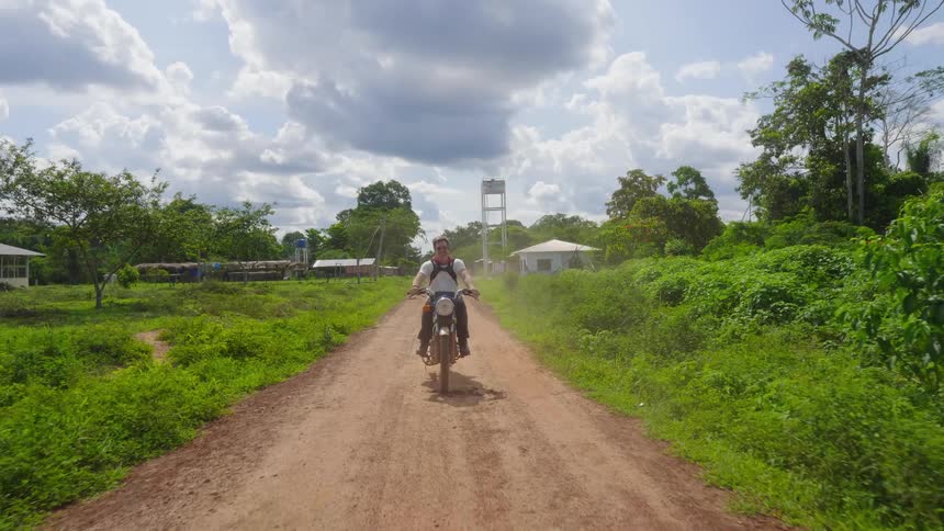 A young man drives a motorcycle down a dirt road in a rural area