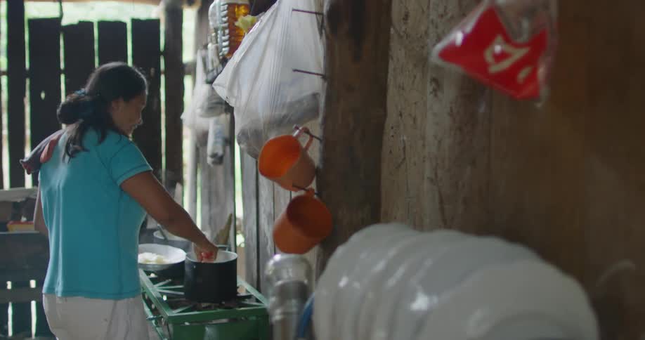 A woman stirs a pot of food while cooking in a small kitchen in a hut