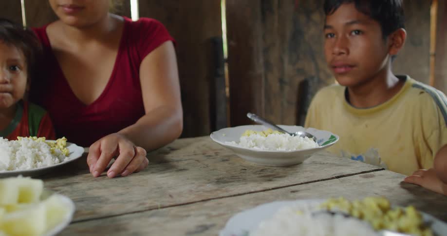 Flies around platefuls of rice on a table where a boy is sitting with his mother and siblings