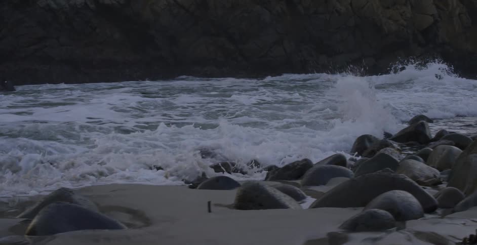 Sea waves crashing on rocks in the sand of a beach next a promontory at sunset