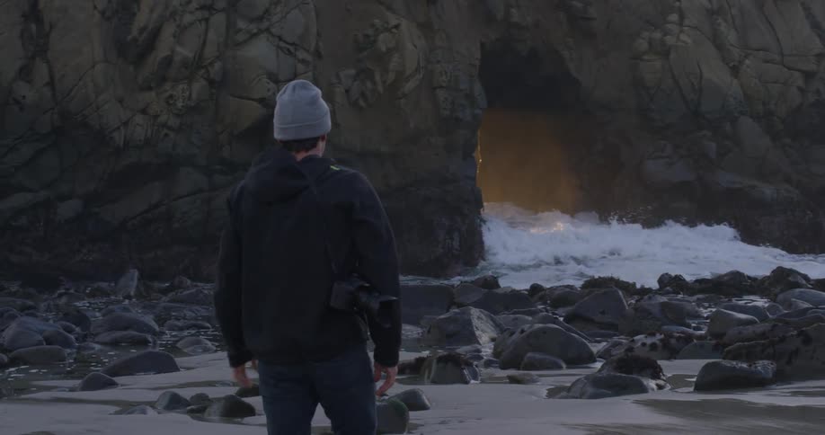 Photographer walks toward the rocks on the beach and photographs sunlight leaking through a promontory