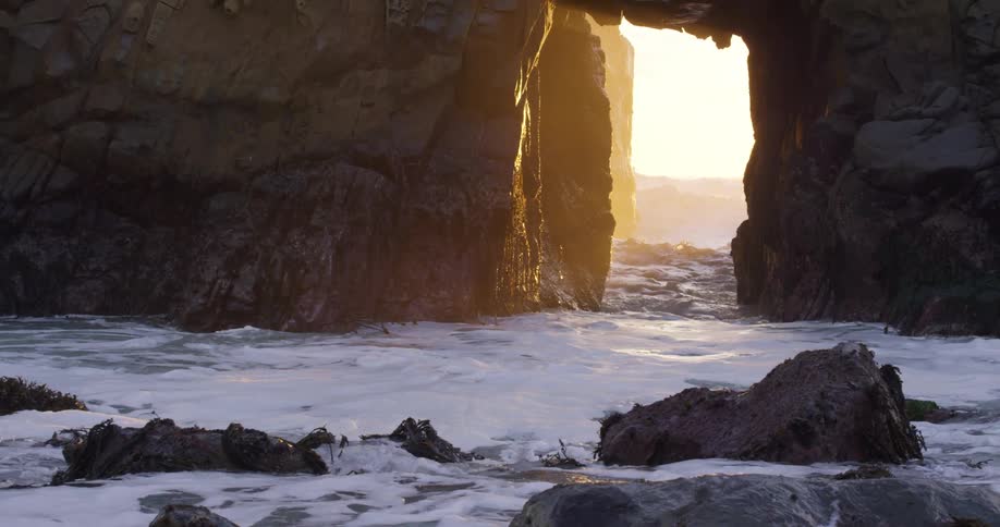 Sunlight shines through a large gap in a rock formation onto a rocky shoreline