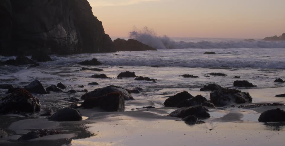Red sky on horizon backs waves crashing against cliff and onto beach