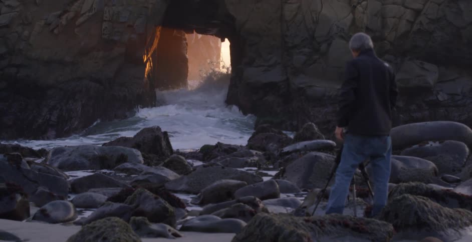 A man stands along a beach with large rocks and waves crashing through a cave
