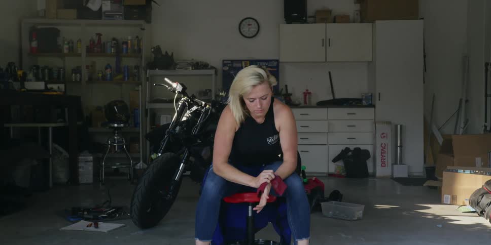 A woman wipes off her hands as she sits in her garage in front of a motorcycle