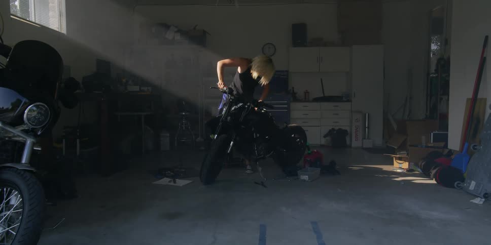 A blonde woman stands behind a motorcycle she is working on in her garage
