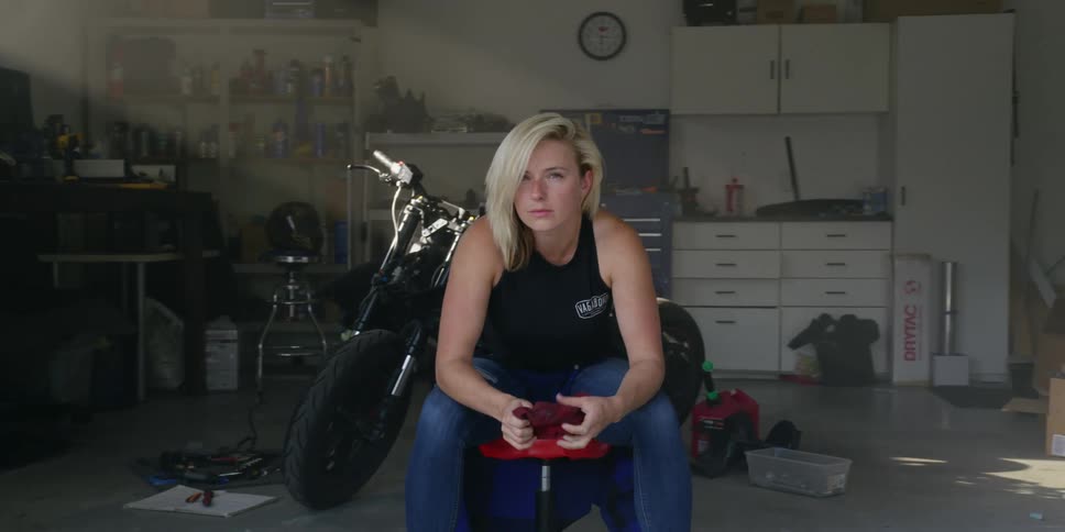 Blonde woman sits on a stool in front of a black motorcycle at the garage
