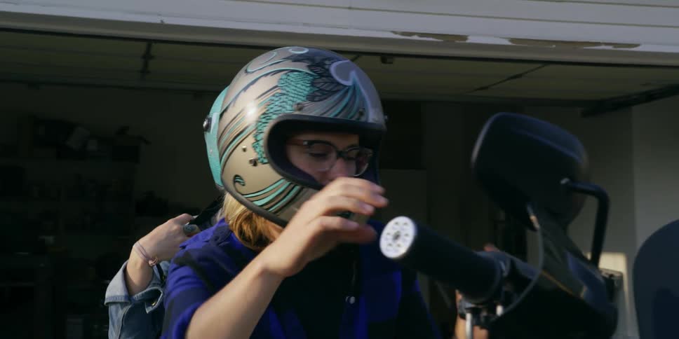 Two women with helmets on a motorcycle in front of a garage with trees in the background