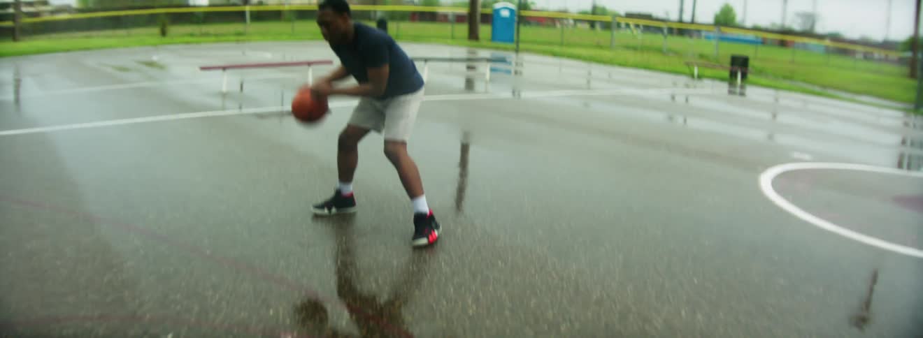 Black man with blue t shirt and black basketball shoes dribbles a basketball in the basketball court on a cloudy day
