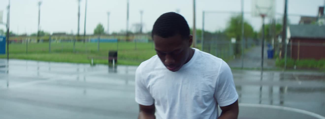 Black man with a white t shirt holds a basketball in the basketball court on a cloudy day