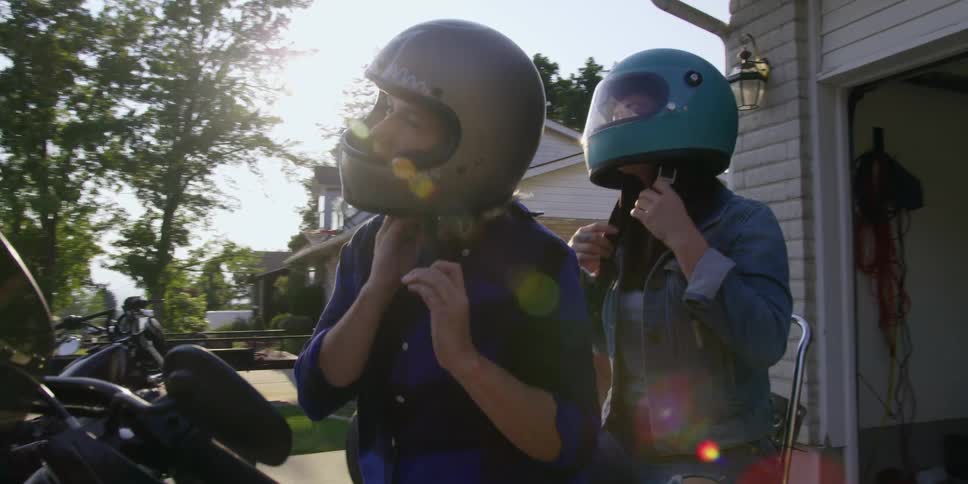 A pair of women put on helmets as they sit on a motorcycle in the driveway of a house