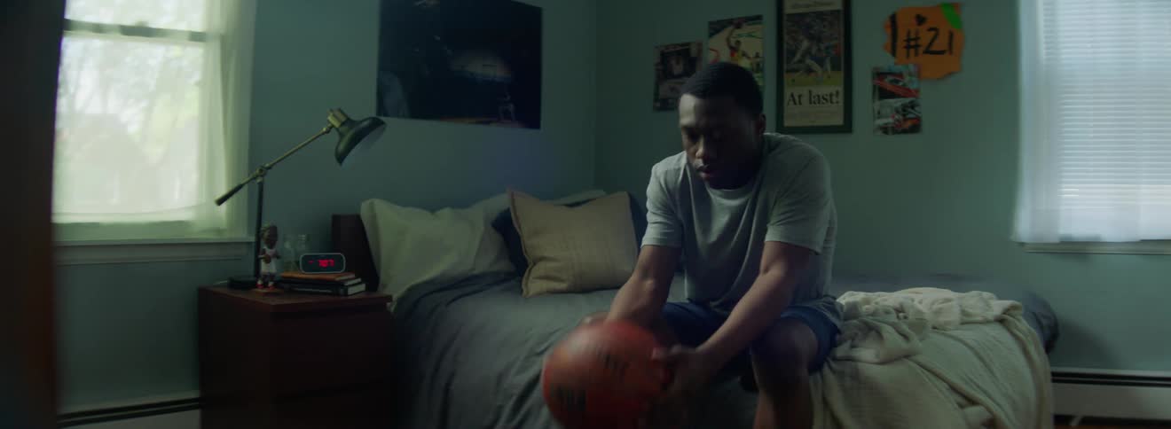 A teenager sits on his bed and picks up a basketball