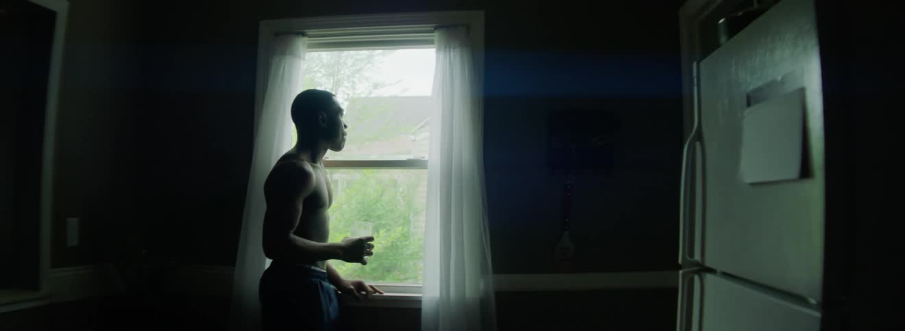 A young black man stands in front of a window in his home and drinks from a glass