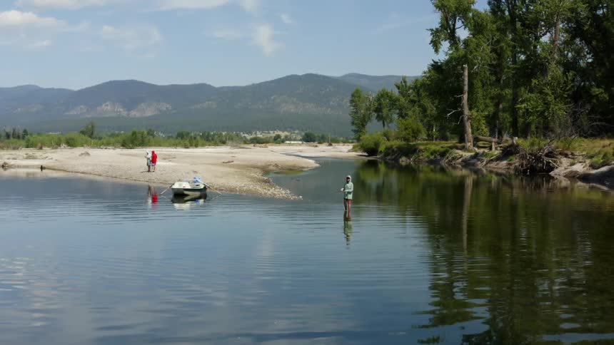 One person stands in the water and fishes as two other people stand on the the shore behind the boat docked there