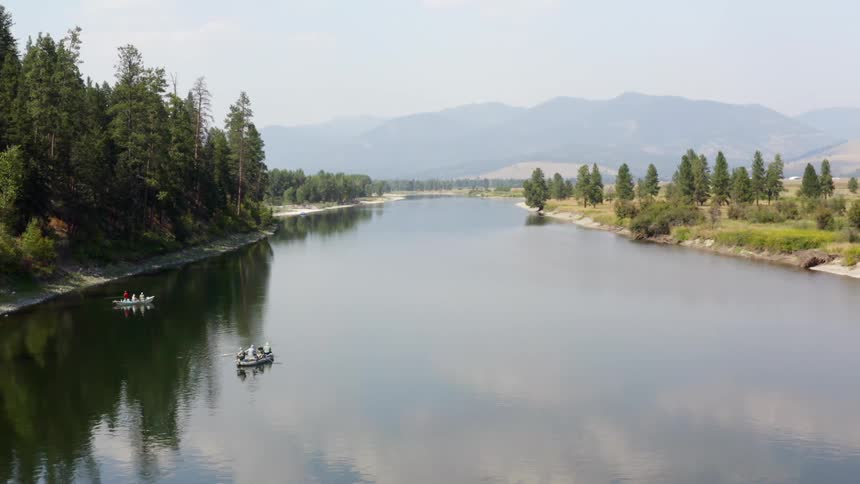 Boats drift near conifer forest covered mountain slope as haze obscures distant mountains