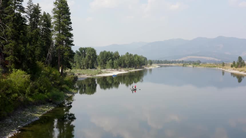 A pair of people paddle a canoe down a river next to a forest