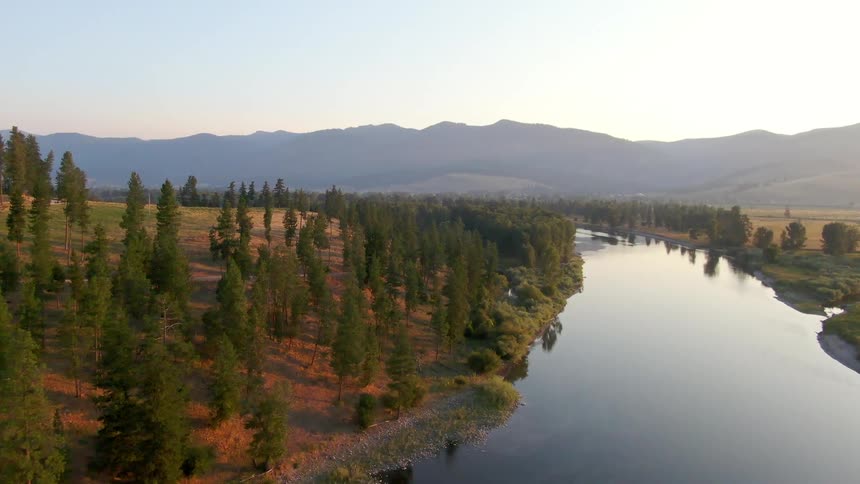 River runs past conifer covered slope turning to parallel misty mountain