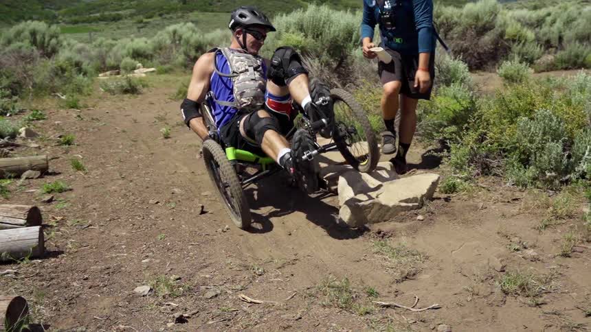 A man helps another man as he bikes over a large rock