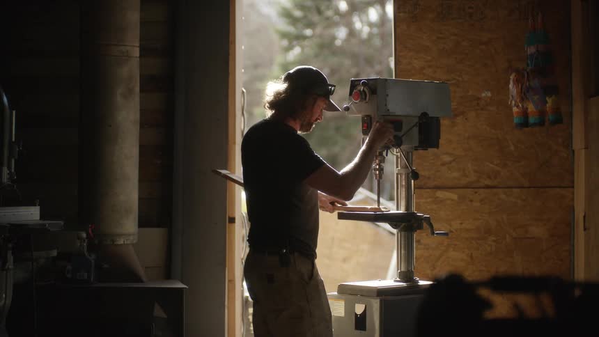 A man drills a hole in wood using a standing drill in his workshop