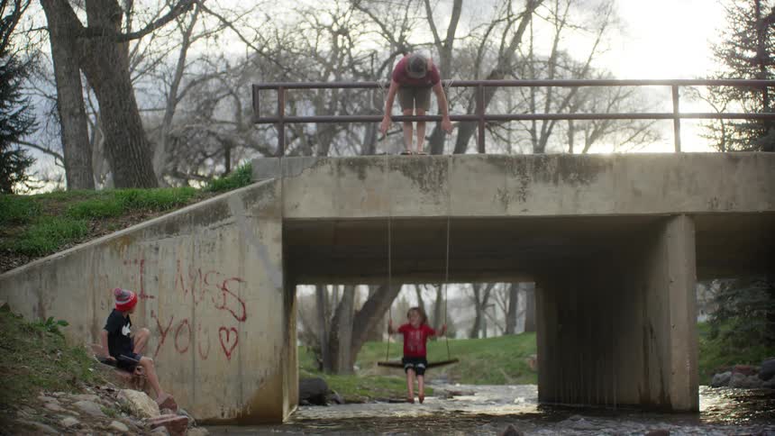 Father swings boy on swing hung off bridge rail over stream