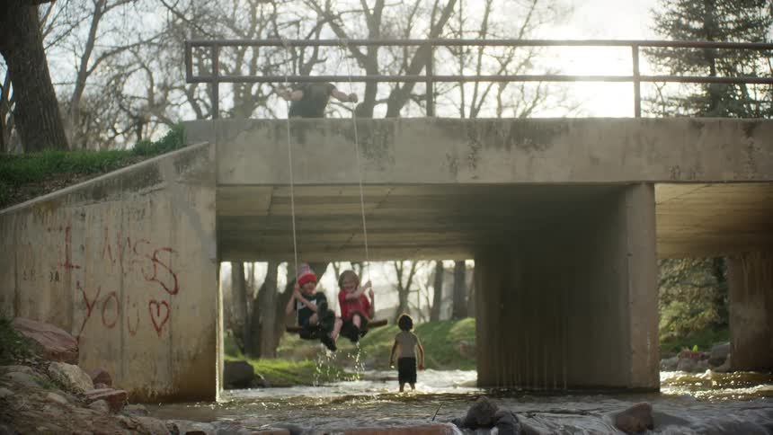 A man pushes a rope on a swing on a bridge while his two kids sit on it and another little boy plays in the river
