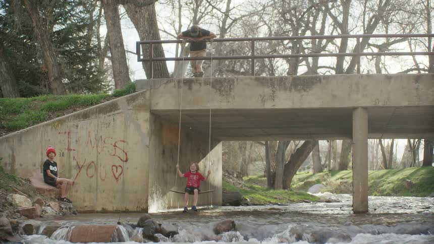 A boy on a swing underneath a bridge with trees and the sky in the background