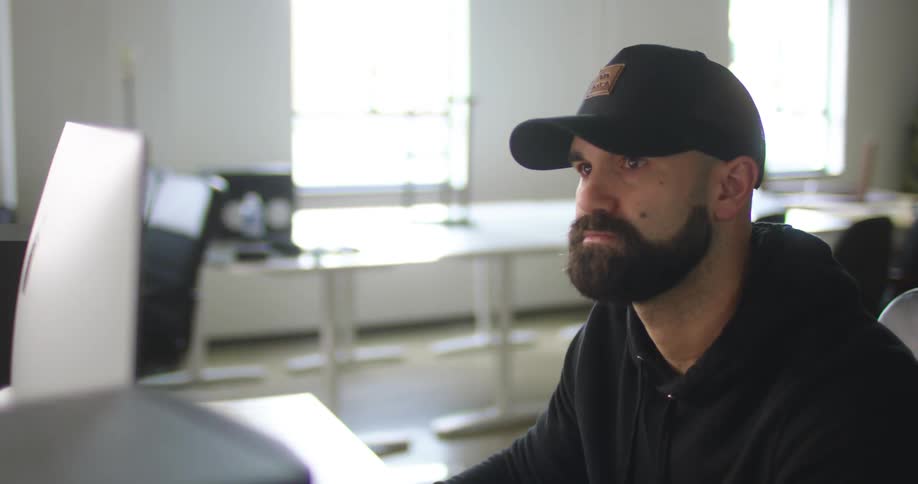 Man wearing hoodie and baseball cap sits in front of computer near empty computer stations