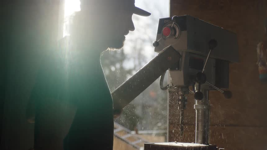 A man uses a large machine drill with a piece of wood in an outdoor workshop