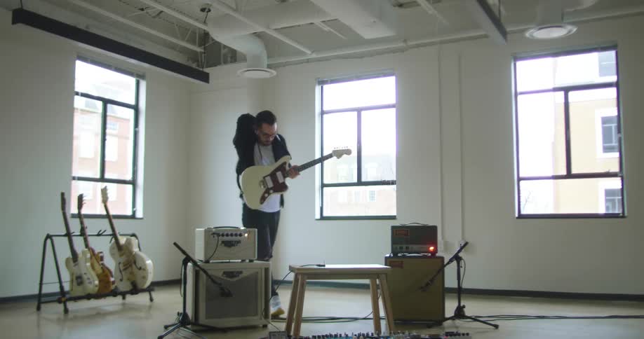 A man walks into a room and picks up a guitar from a rack plugs it in and turns on the amp