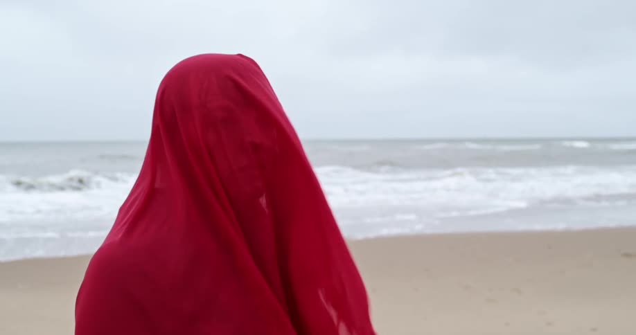 A woman with a sheer red scarf covering her head and face stands with her back to the ocean waves