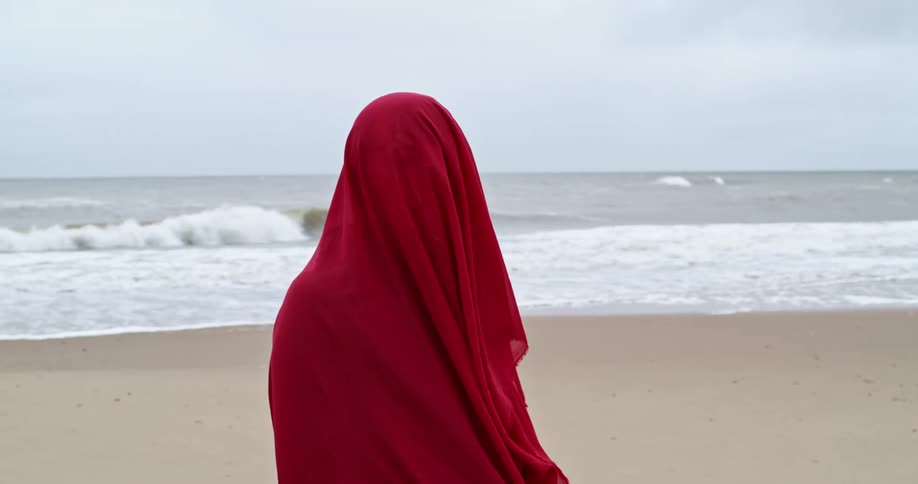 A woman covered up with a cloak stands with her back to the water